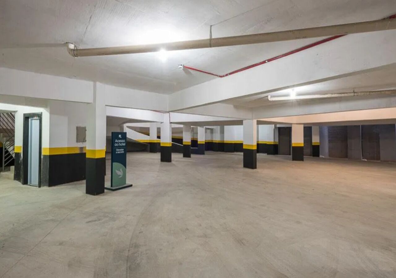 Amplo estacionamento coberto com diversas vagas, elevador visível e sinalização indicando acesso a um hotel.