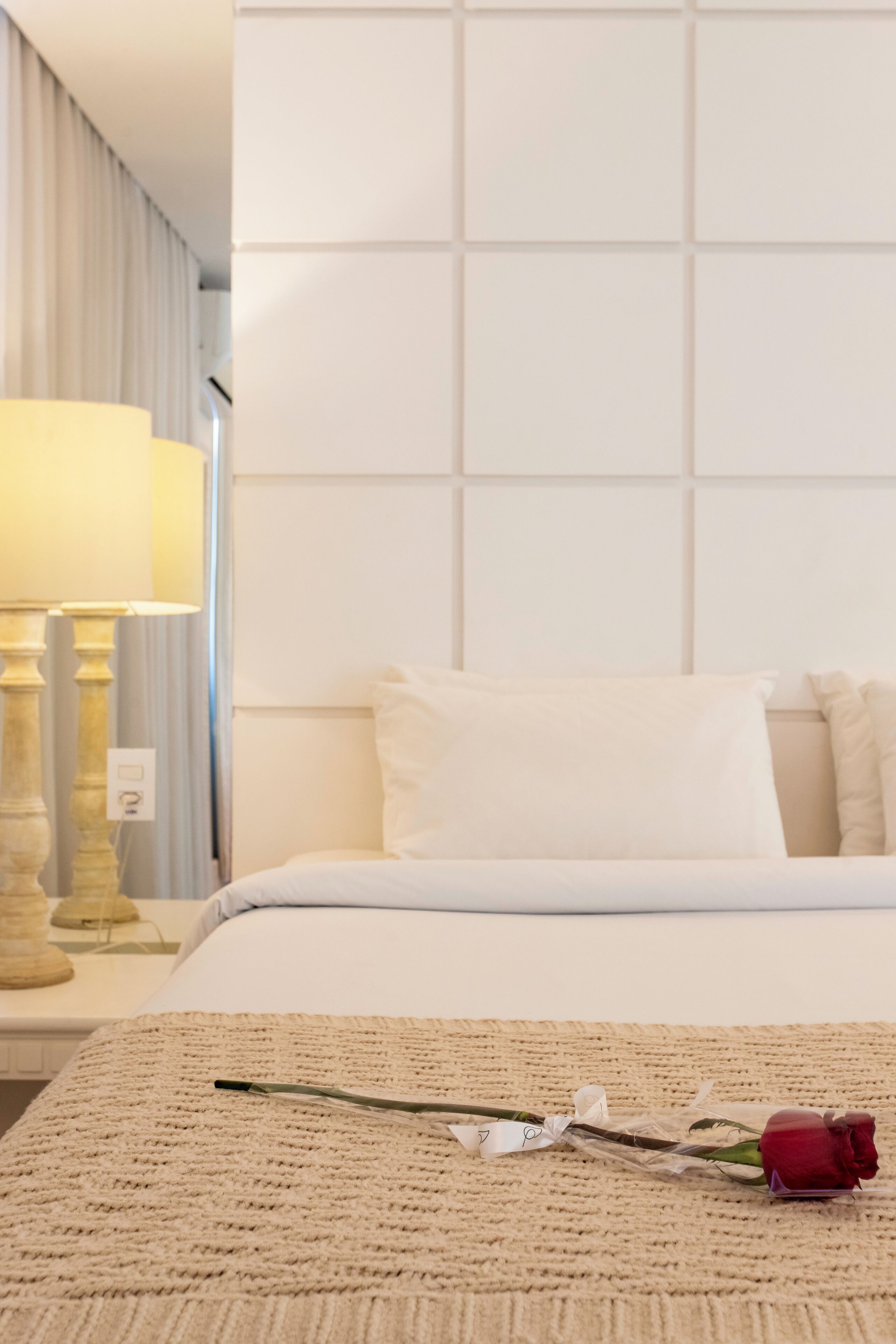 Ein Hotelzimmer mit einem ordentlich bezogenen Bett, einer Nachttischlampe und einer einzelnen roten Rose auf dem Bett.