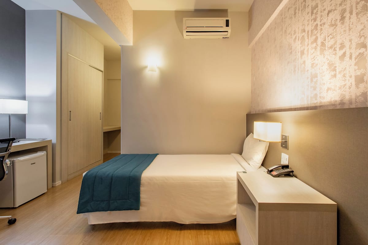 Hotelkamer met een bed, nachtkastje, lamp, telefoon en bureau met stoel en een kleine koelkast.