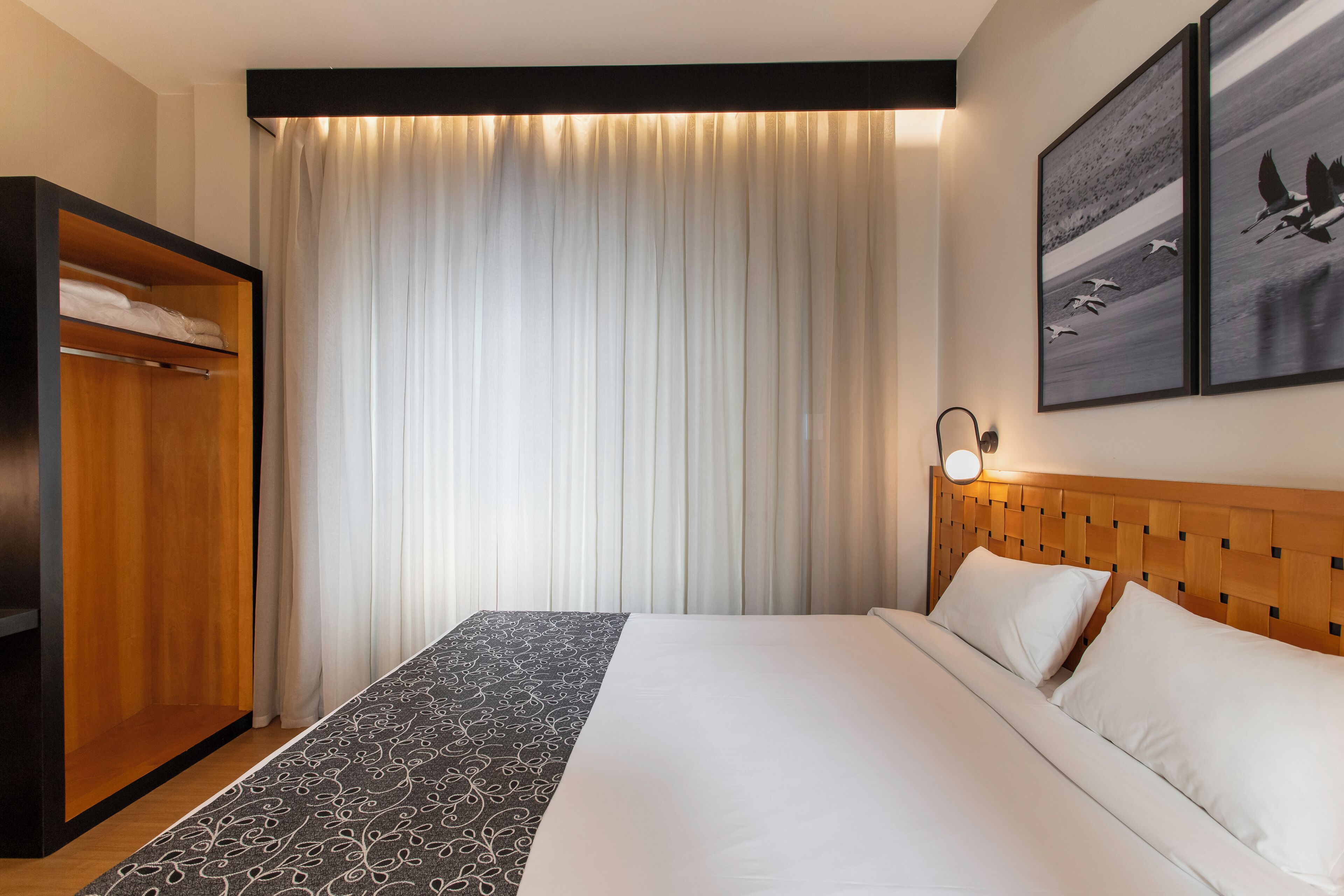 A hotel room with a large bed, a wardrobe, and a view of the outdoors through sheer curtains.