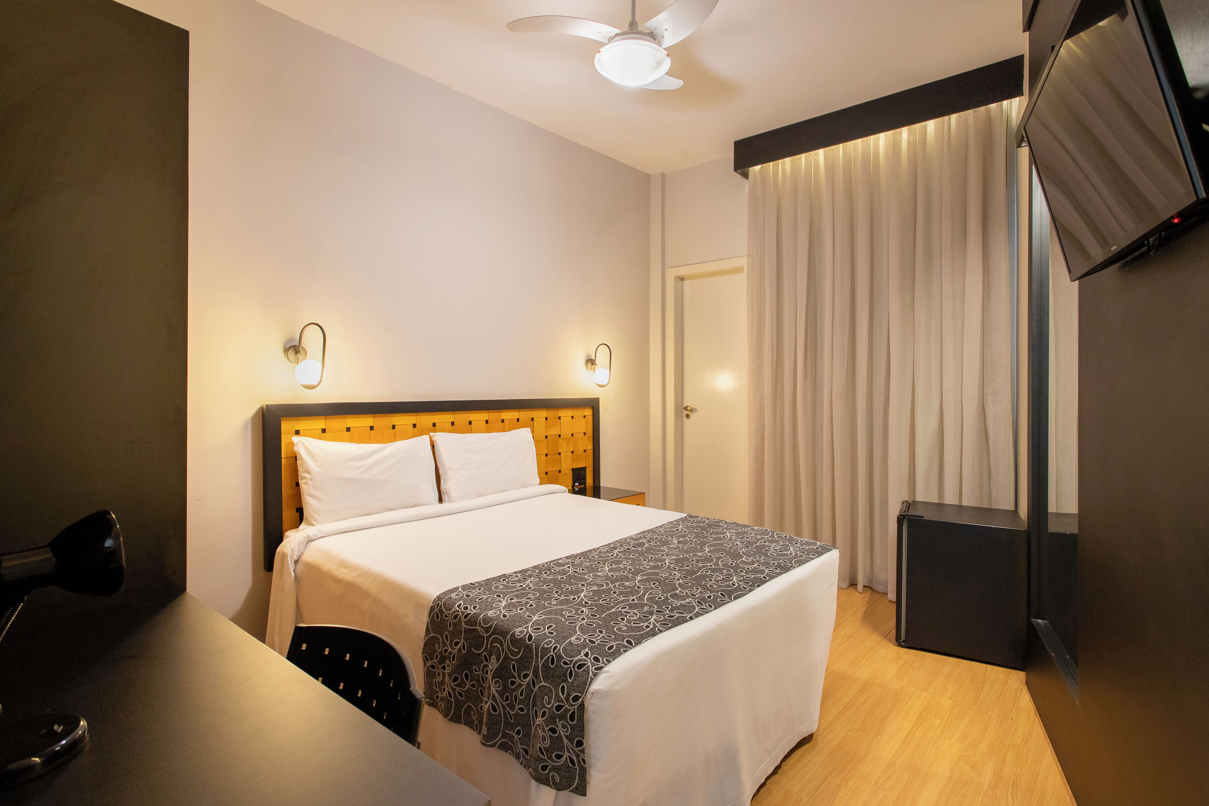 Quarto de hotel com cama, abajures, televisão de tela plana e ventoinha no teto.