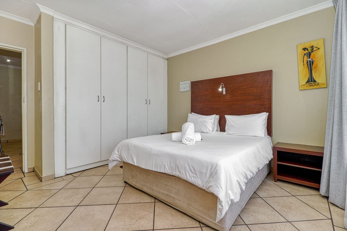 A hotel room with a bed, white curtains, a wooden headboard, and a painting on the wall.