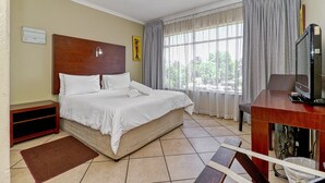A hotel room with a bed, a desk with a television, a window with curtains, and a wall-mounted air conditioning unit. - BM Gardens Apartment Hotel (Germiston)