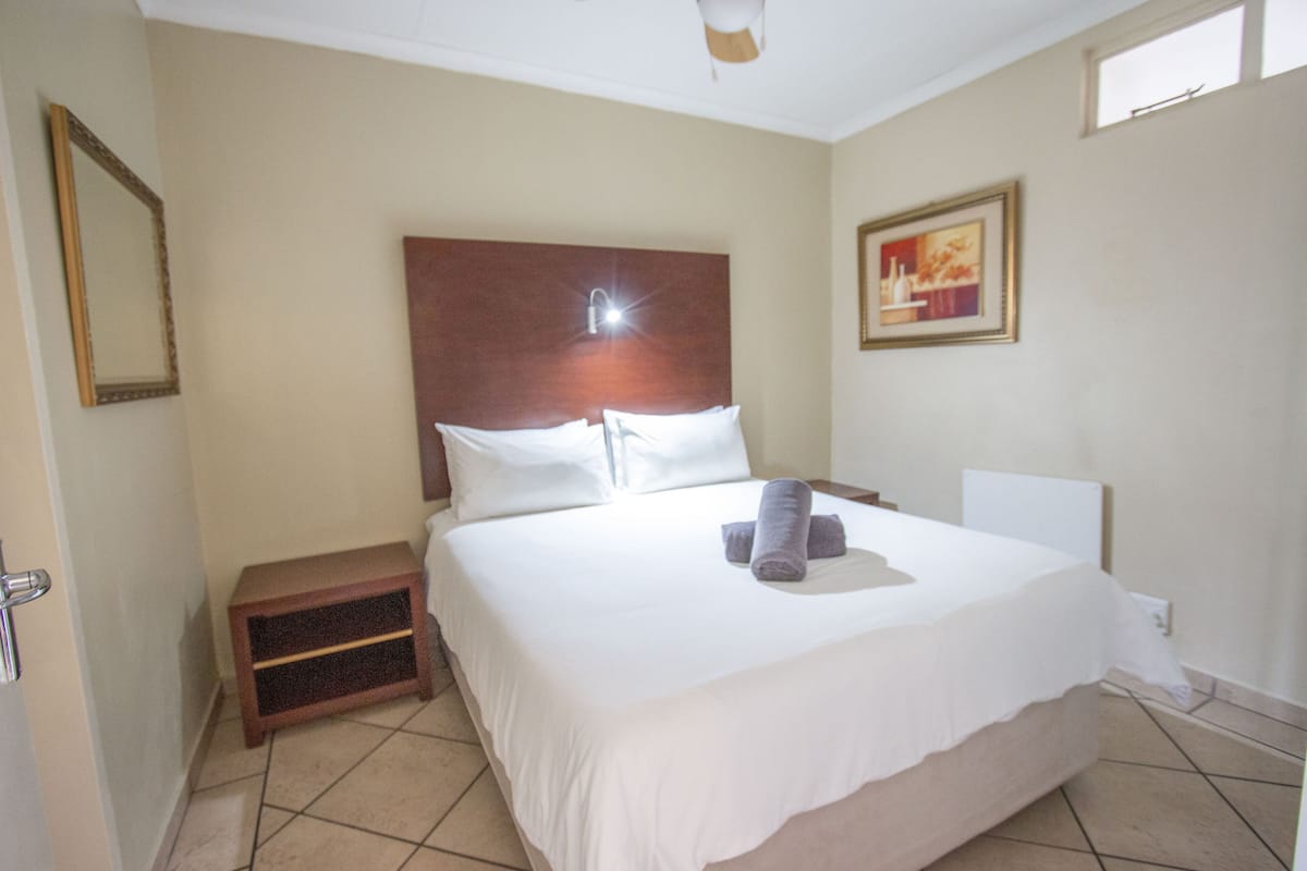A hotel room with a large bed, a nightstand, a ceiling fan, a framed picture on the wall, and a window.