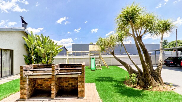 A brick barbecue grill in a grassy yard with a playground and a palm tree. - BM Gardens Apartment Hotel (Germiston)