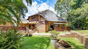 Een bungalow met rieten dak, houten balkon en een verzorgde tuin met palmbomen en een grote rots als middelpunt.