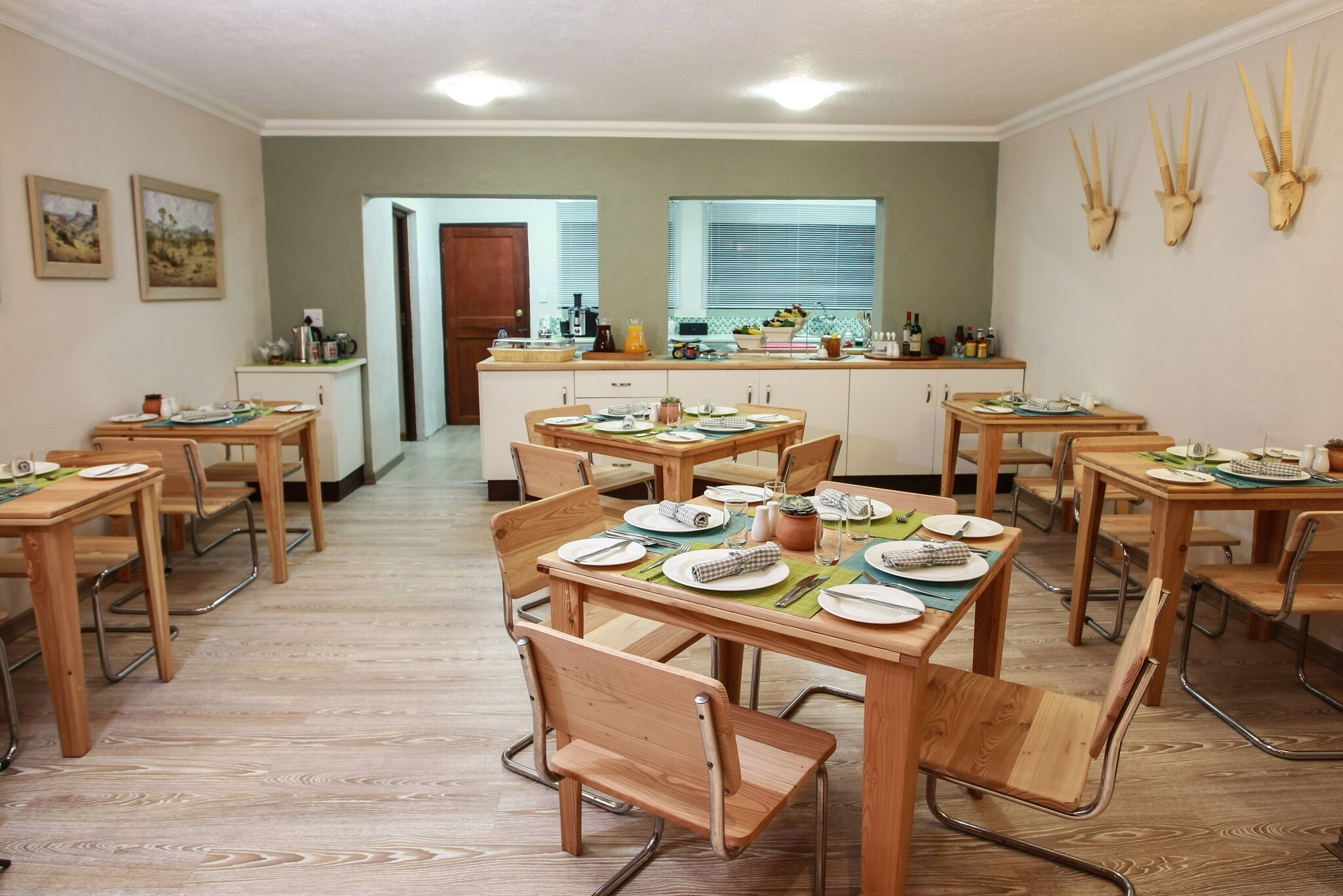 A dining area with multiple wooden tables and chairs, a kitchen in the background, and decorative antelope heads on the wall.