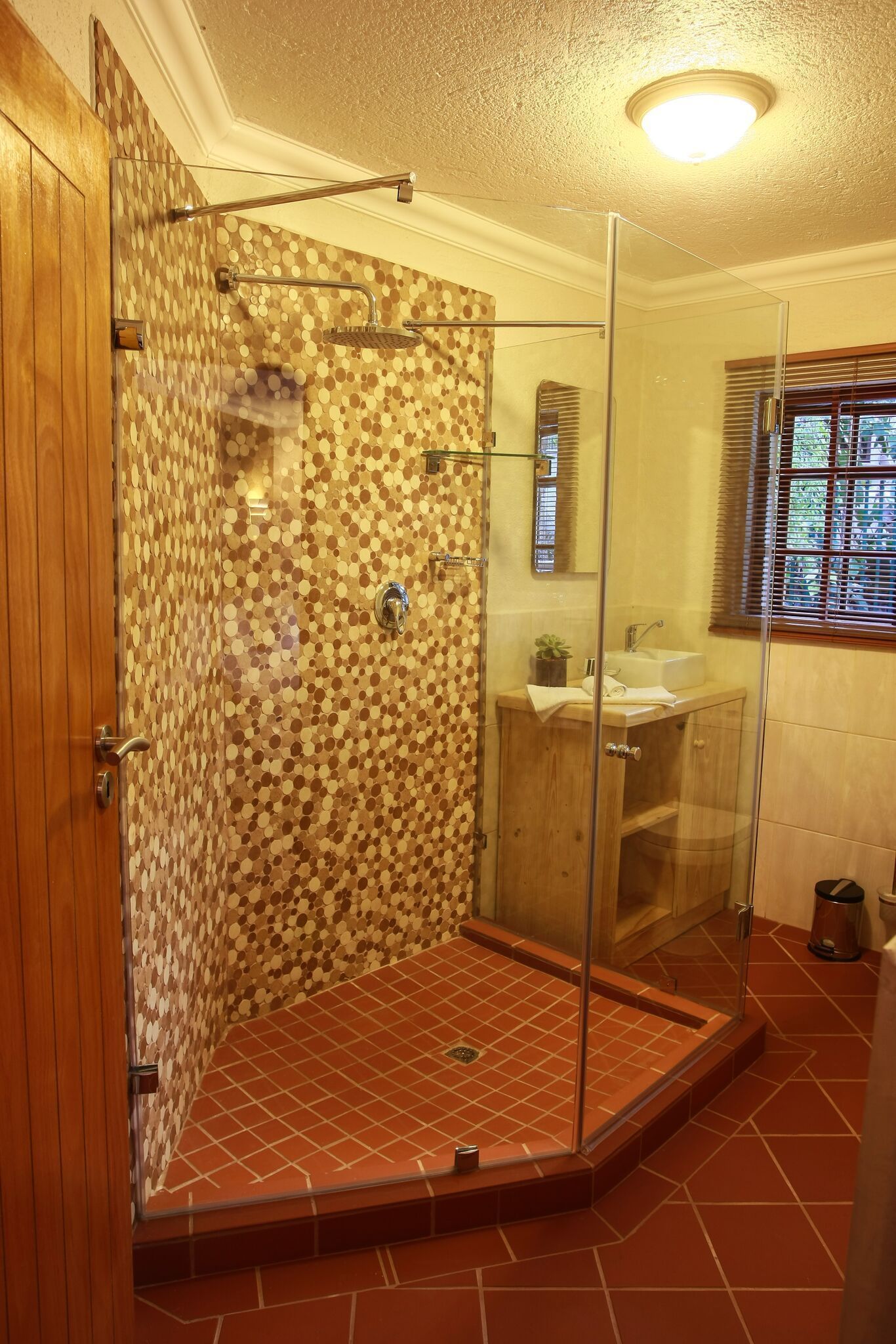 Une douche avec une porte en verre, un meuble-lavabo en bois et une fenêtre avec des stores.
