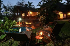 Un espace aménagé au bord de la piscine, avec des chaises longues et un jacuzzi, entouré d’une végétation luxuriante et d’une maison au toit de chaume.
