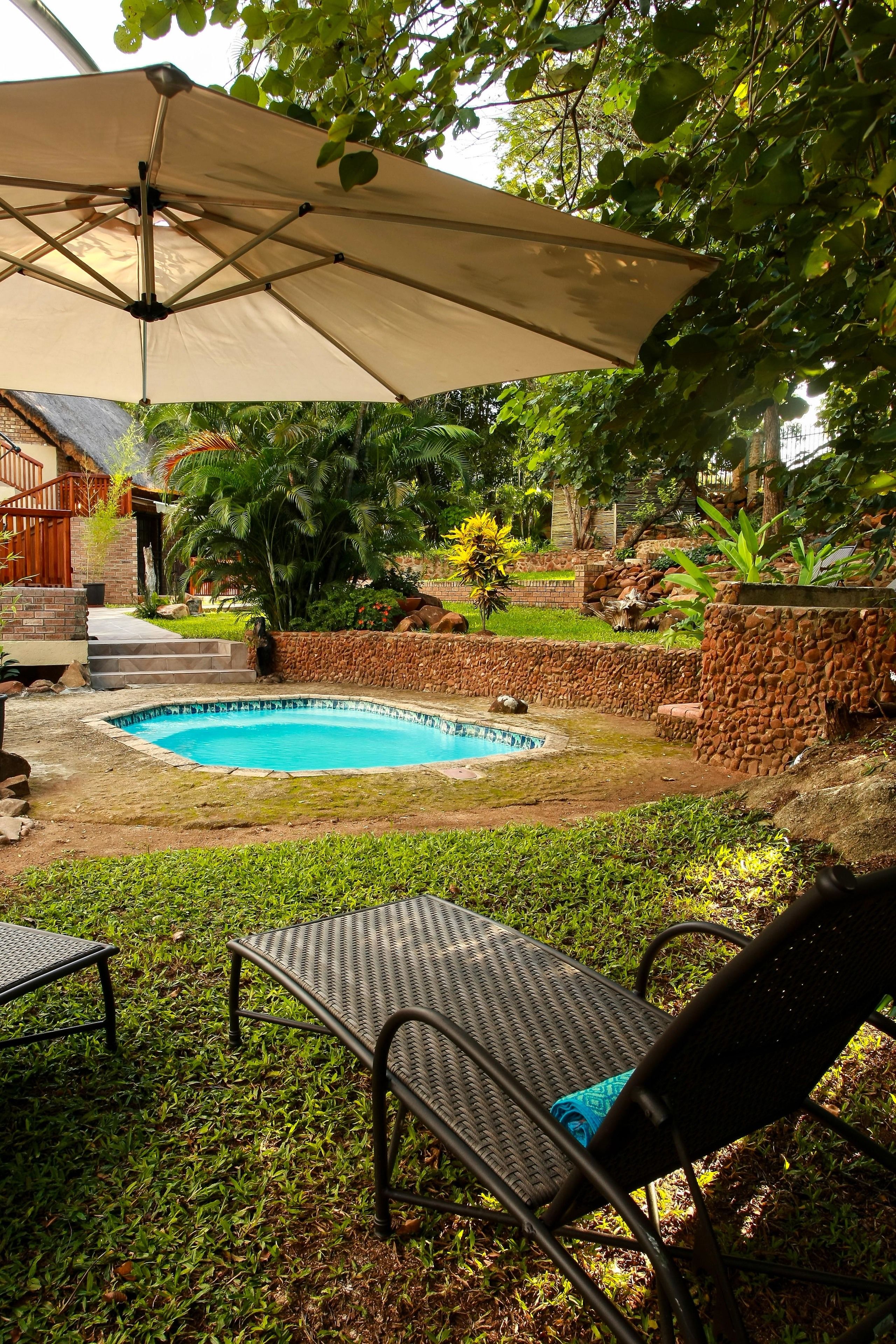 Un jardin avec une piscine, entouré d’un mur en pierre, et comprenant un patio couvert avec des chaises.