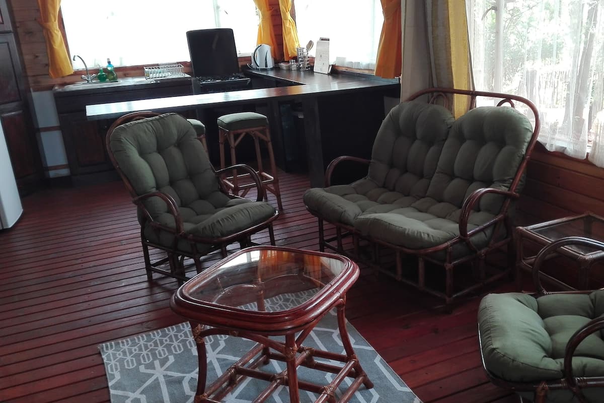 A cozy room with wicker furniture, a wooden floor, a kitchen area with a stove and sink, and large windows with curtains.
