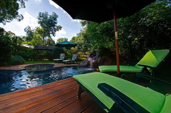 Une terrasse en bois avec un jacuzzi, des fauteuils de jardin verts et une cascade décorative.