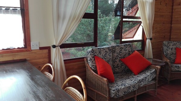 Interior de una cabaña de madera con mobiliario de mimbre, una mesa de comedor y ventanas amplias con cortinas.