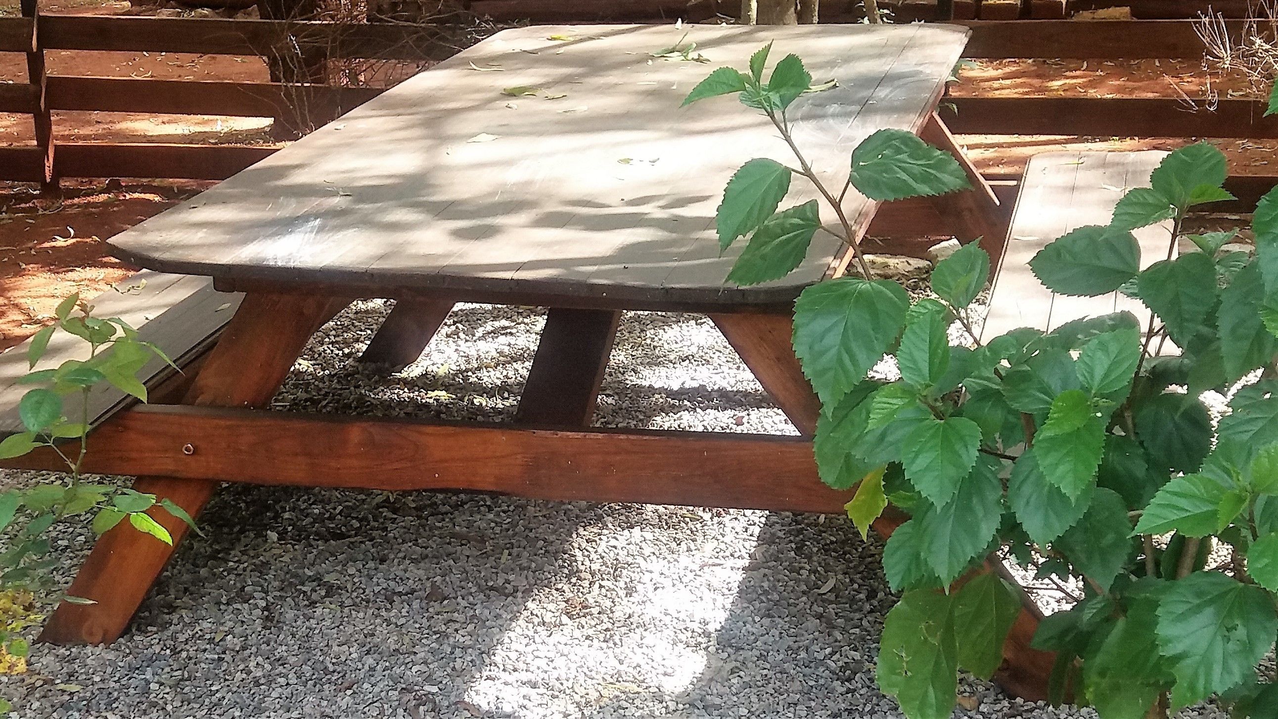 A wooden picnic table with a concrete top, surrounded by gravel and greenery.