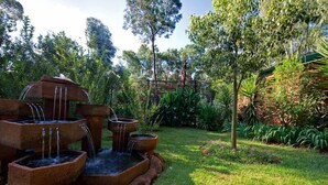 A garden with a multi-tiered stone fountain surrounded by lush greenery and trees. - Woodland Gardens (Magaliesberg)