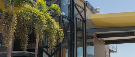 Un edificio moderno con ascensor de vidrio, palmeras y una zona de estacionamiento techada.