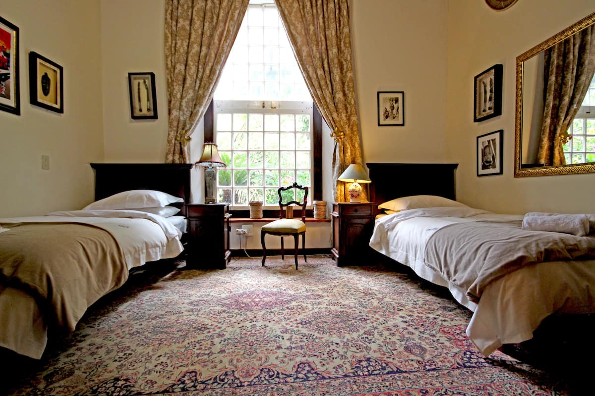 A room with two beds, a desk, and a chair. There are curtains, framed pictures on the walls, and a window with a view of greenery.