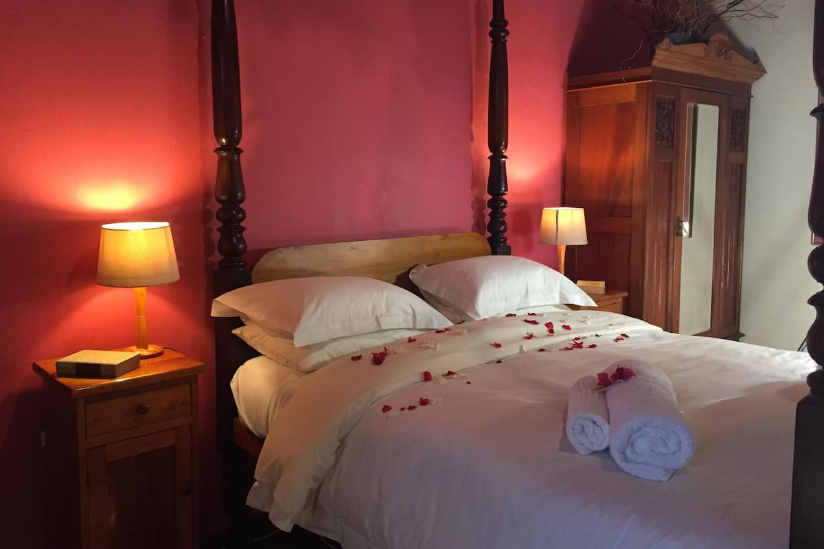 A four-poster bed with white linens and rose petals, two bedside lamps, and a wooden nightstand with a lamp.