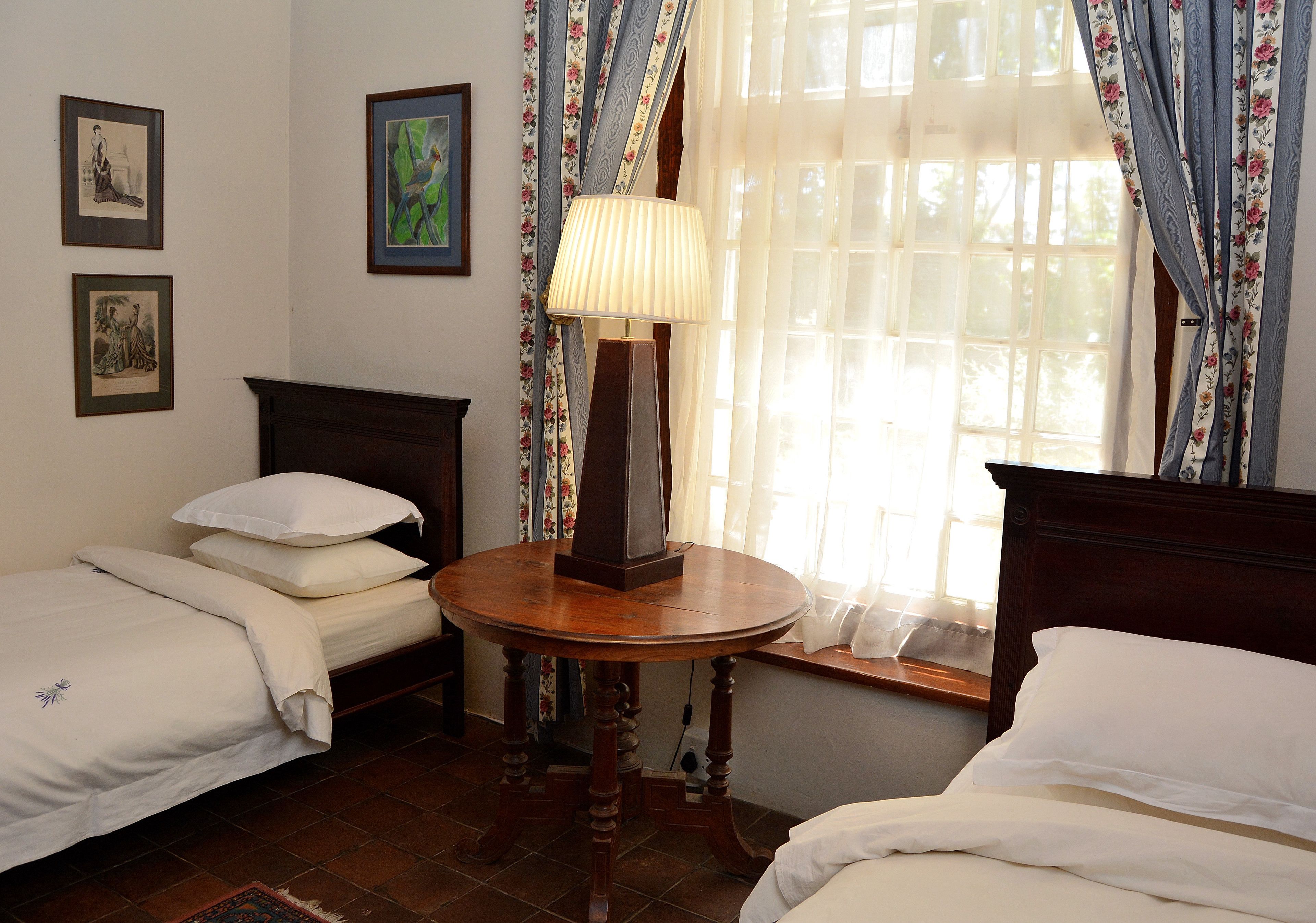 A room with two single beds, a wooden table with a lamp, and a window with floral curtains.