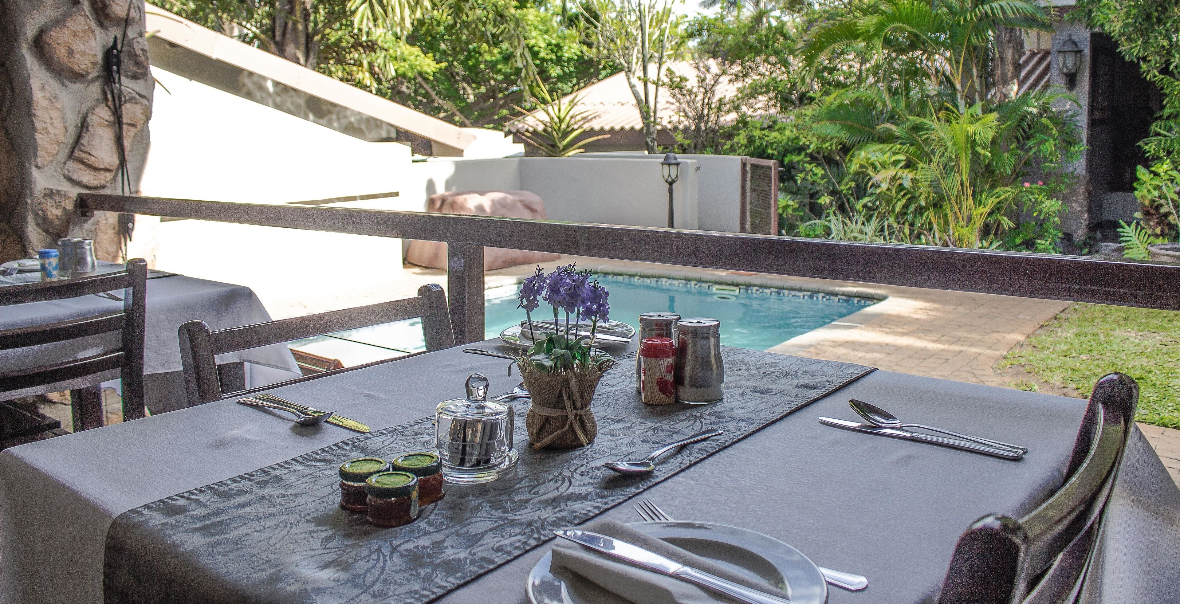A dining table set outdoors with a pool view, a stone wall, and lush greenery.