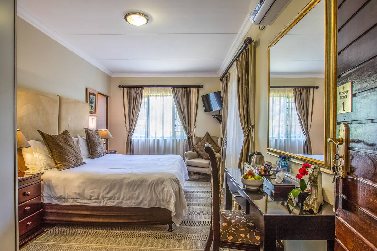 A well-lit hotel room with a large bed, a desk with a mirror, a chair, and a small table with a vase of flowers.