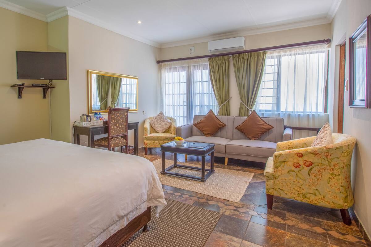 A hotel room with a bed, a sofa, two armchairs, a small table, a desk with a chair, a television, and a window with curtains.