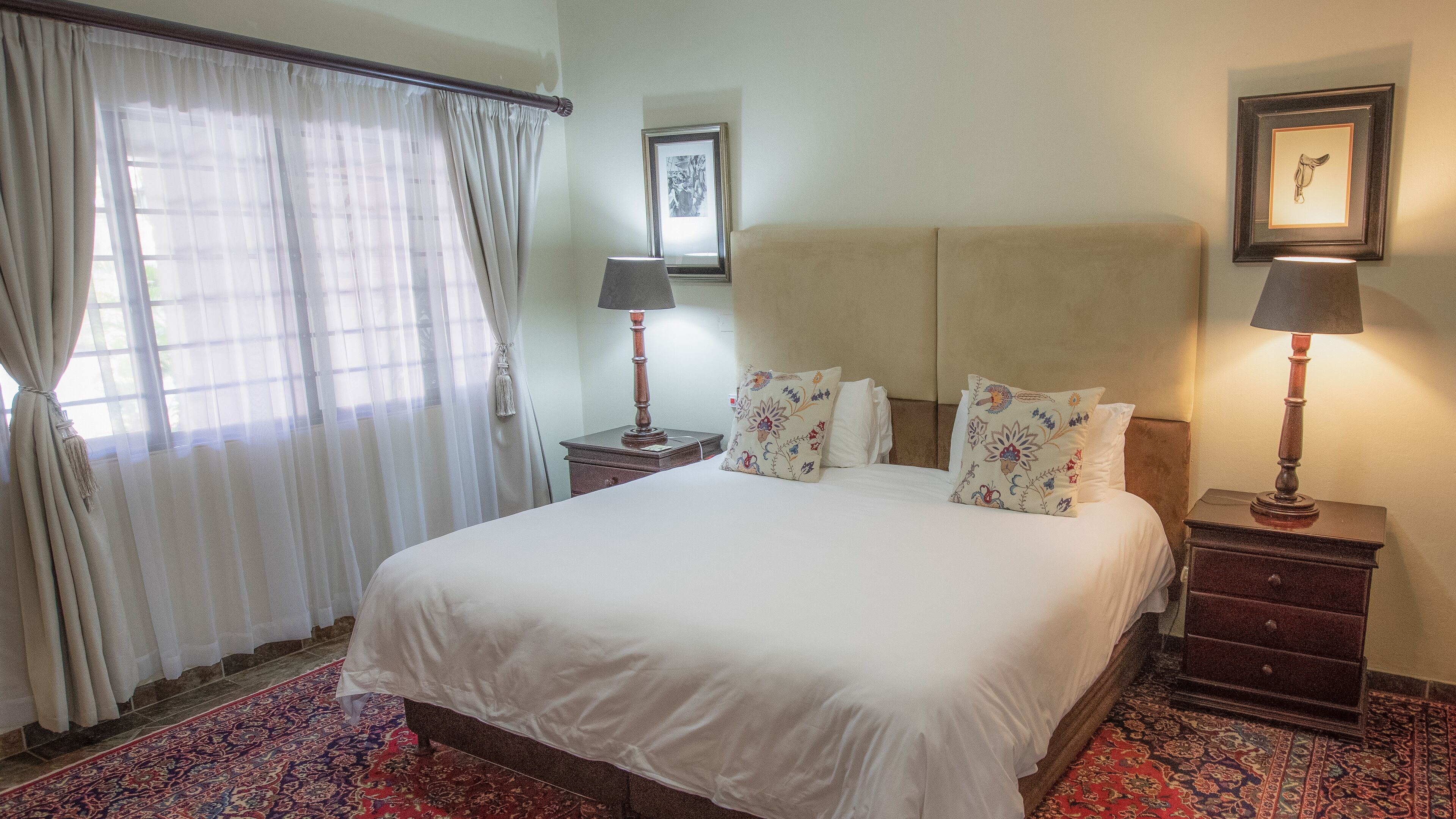 A hotel room with a large bed, two bedside tables with lamps, a window with sheer curtains, and a framed picture on the wall.