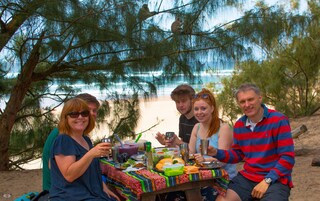 Un grupo de personas cenando en una mesa al aire libre con vista a la playa y árboles.