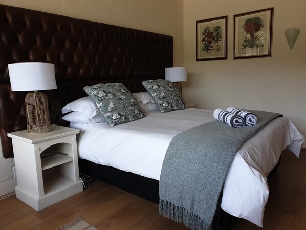 A neatly made bed with a grey blanket, two matching pillows, and a decorative lamp on a white nightstand. - Leeuwenzee Guest House (Cape Town)