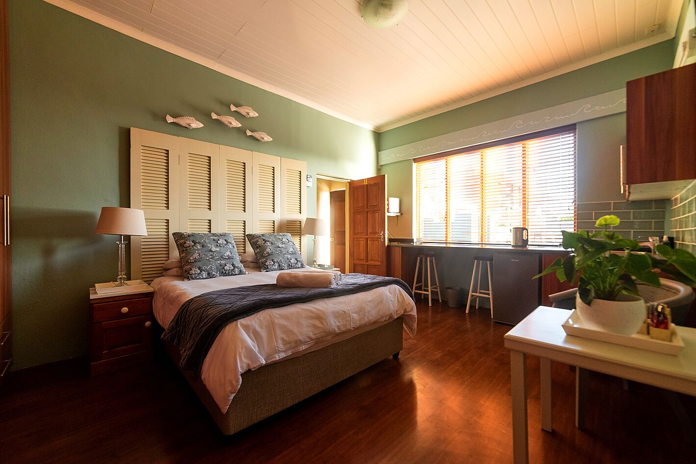 Ein Schlafzimmer mit Bett, Nachttischen, einem kleinen Hocker, einer Pflanze und einer Küchenzeile. Vor dem Fenster sind Jalousien.