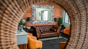 A cozy living room with a brown leather sofa, a round mirror, and a brick wall. - Leeuwenzee Guest House (Cape Town)