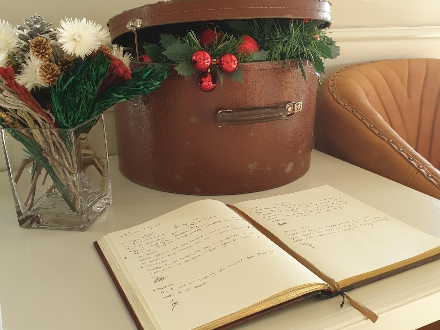 An open book with handwritten notes, a clear glass vase with dried flowers, and a brown leather bag with red ornaments.