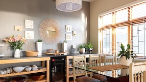 A compact kitchen with a dishwasher, wooden shelves, a glass dining table, and a dining area with chairs. - Leeuwenzee Guest House (Cape Town)