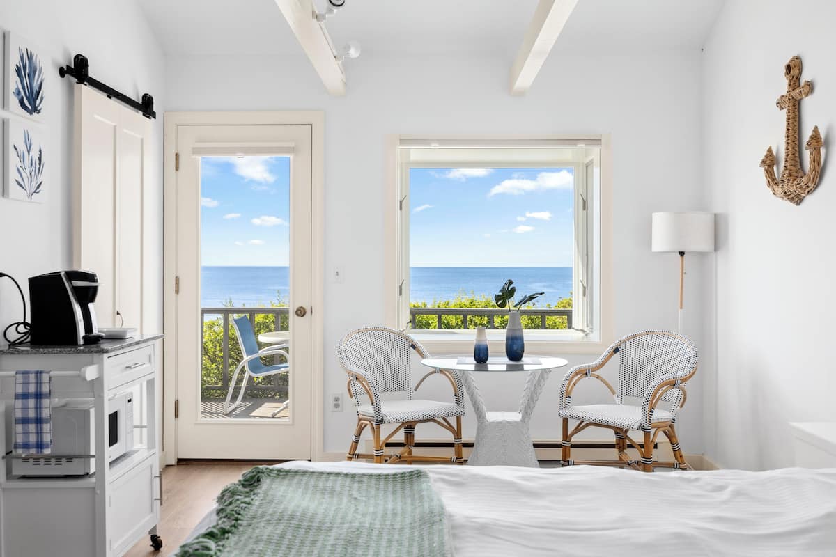 A room with a bed, two wicker chairs, a small table, a coffee maker, and a view of the sea through a window.