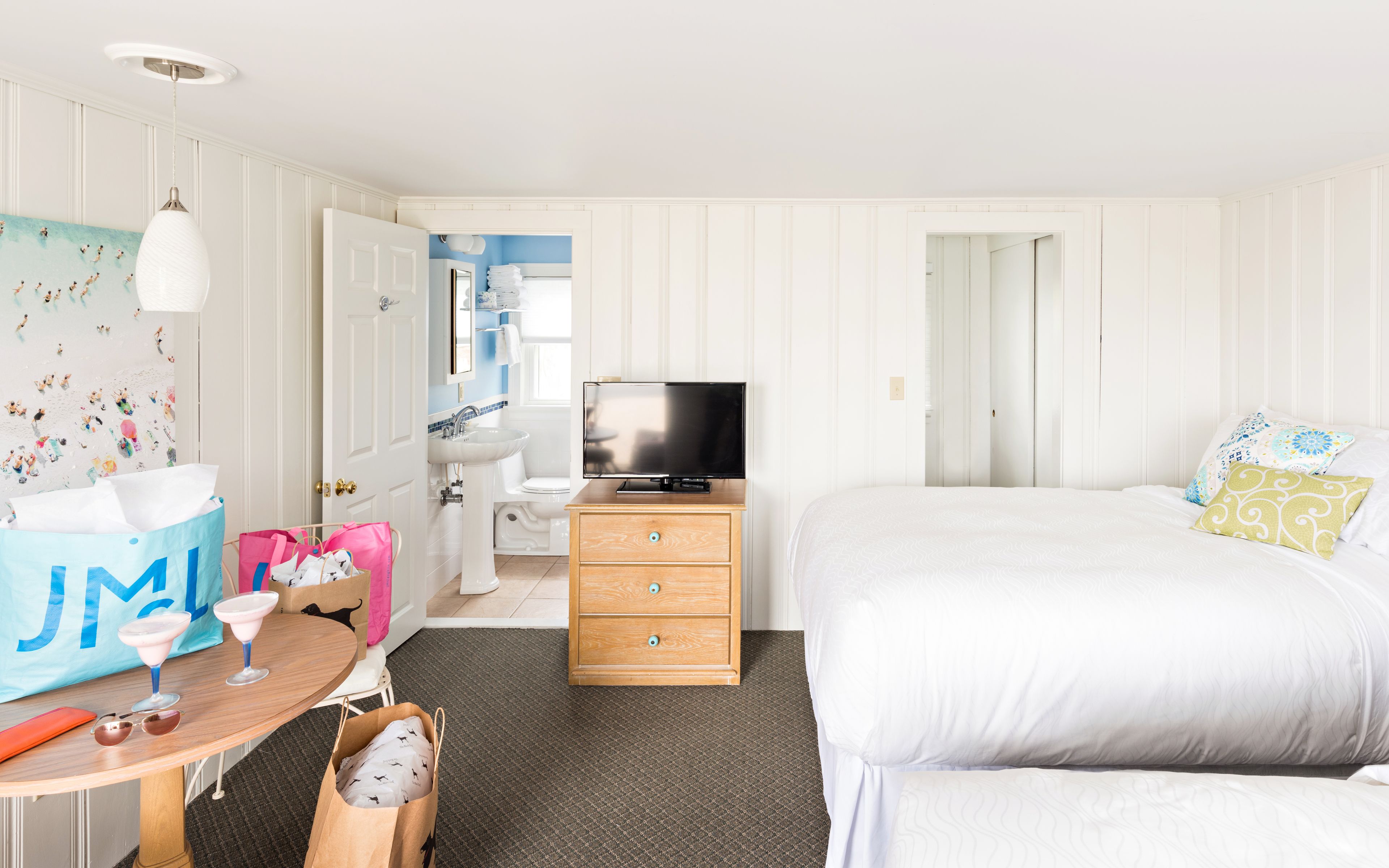 Habitación de hotel con cama, televisor, cómoda de madera, baño con lavamanos e inodoro, y una mesa con bolso y gafas de sol.