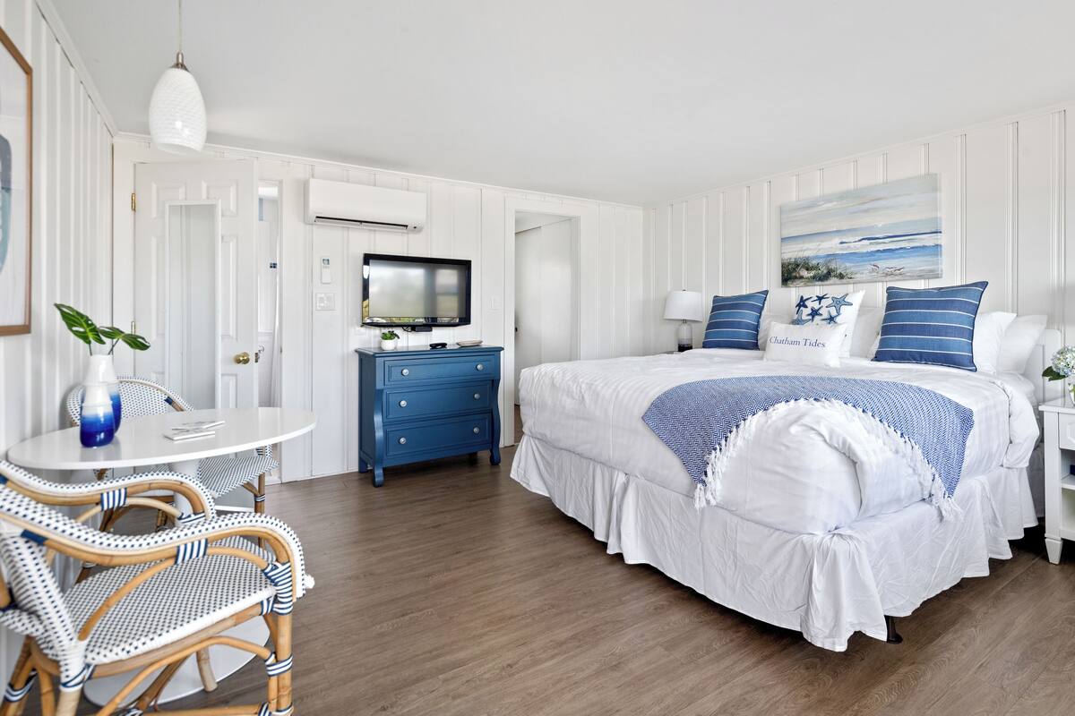 A hotel room with a bed, a TV, a desk with a chair, and a window with a view of the beach.