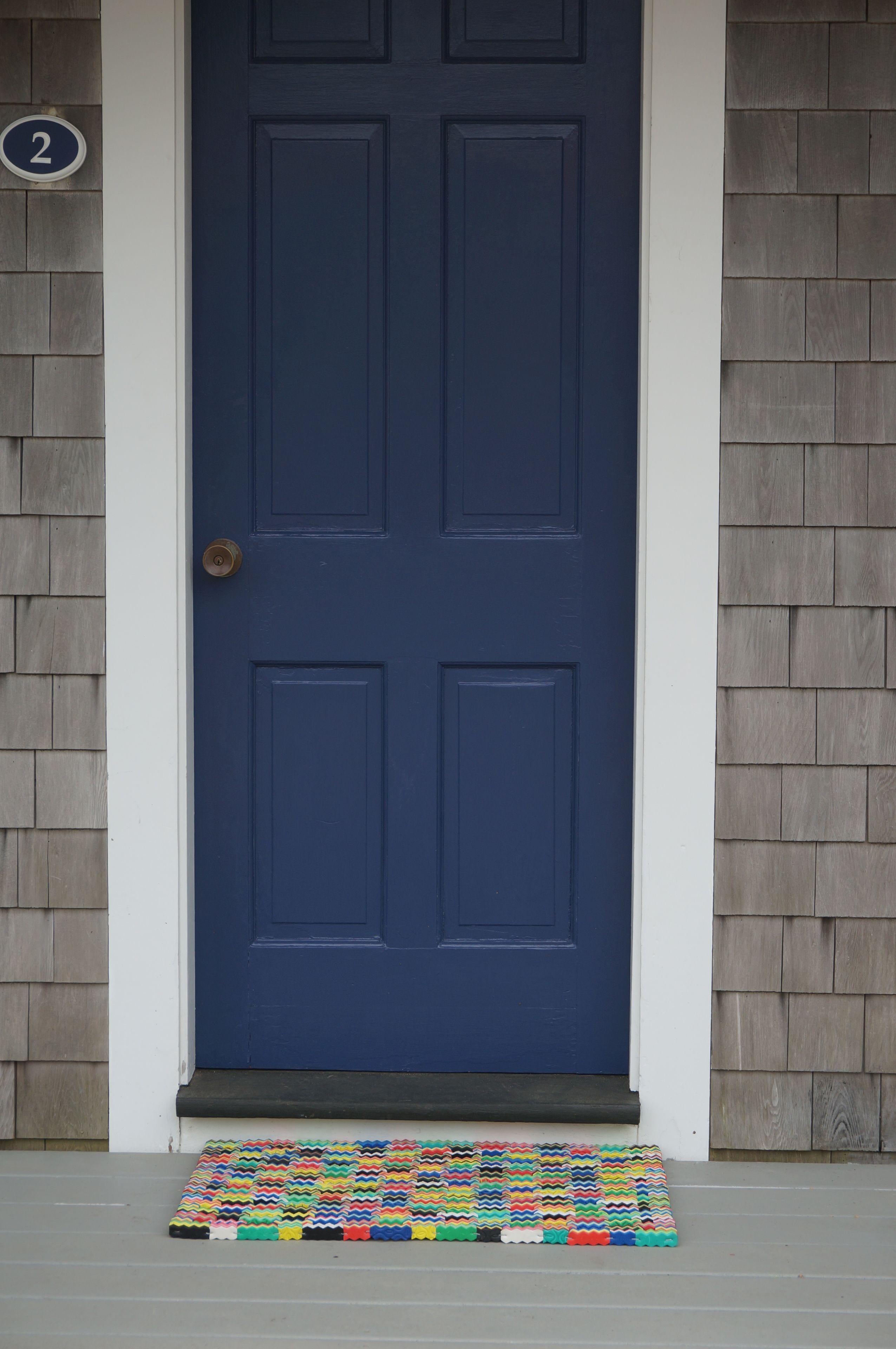 A dark blue door with a number '2' sign above it, a colorful mat at the entrance, and a wooden shingle wall.