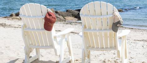 Deux chaises Adirondack blanches sur une plage de sable, avec une côte rocheuse et l’océan en arrière-plan.
