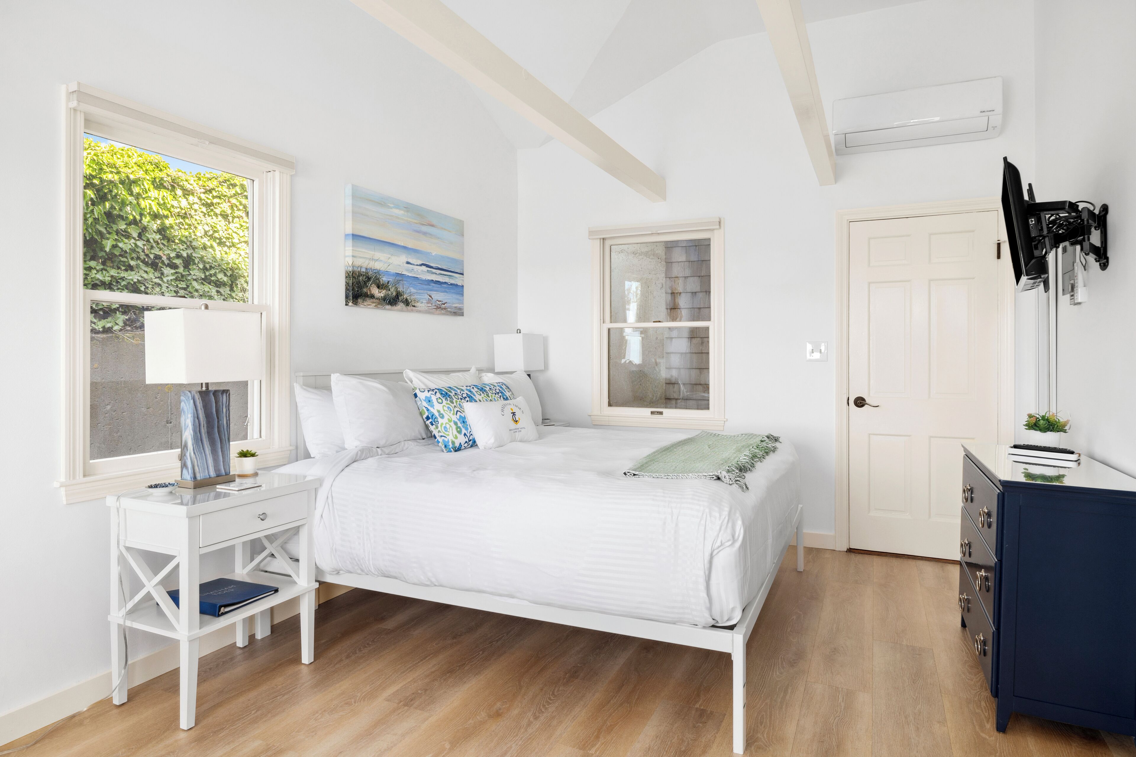 A bedroom with a bed, bedside tables, a desk, a dresser, and a window with a view of greenery.