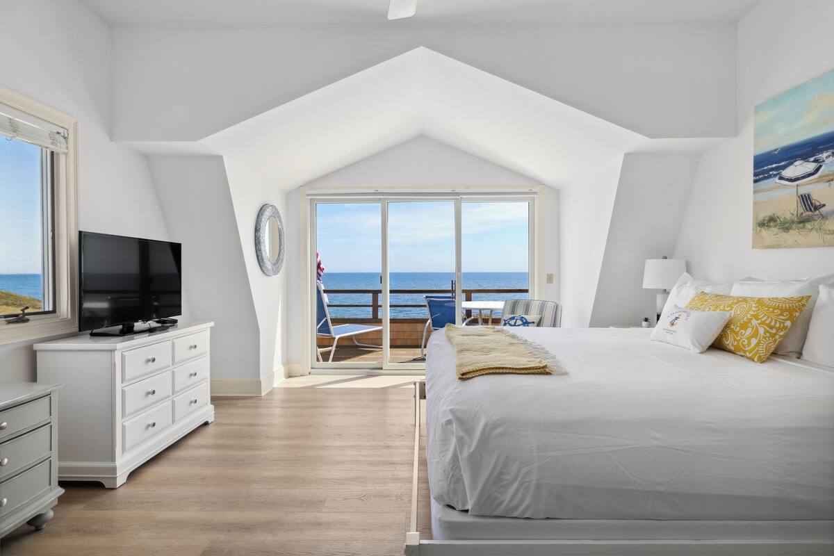 A bedroom with a large bed, a TV, a dresser, and a balcony with a view of the sea.