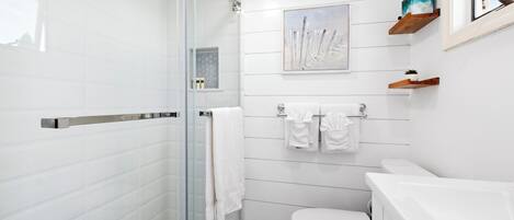 Ein modernes Badezimmer mit Duschkabine aus Glas, weiß gefliesten Wänden, einer weißen Toilette und einem aufgehängten Bild.