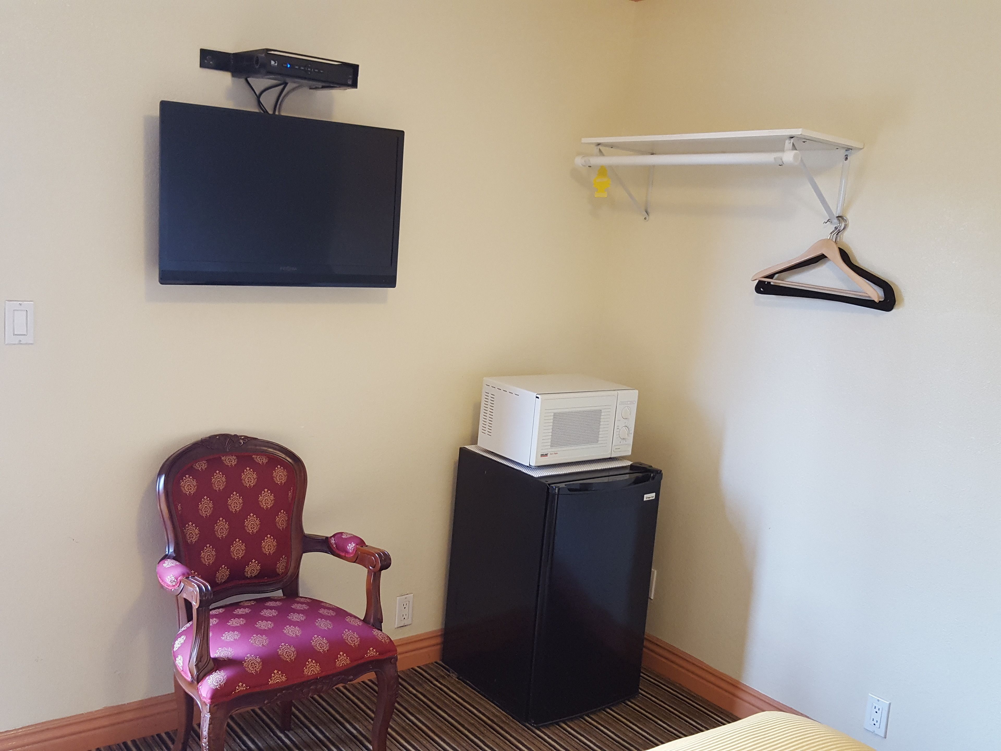 A room with a television mounted on the wall, a small refrigerator, a chair, and a shelf with a hanger.