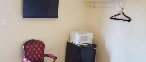 A room with a television mounted on the wall, a small refrigerator, a chair, and a shelf with a hanger.