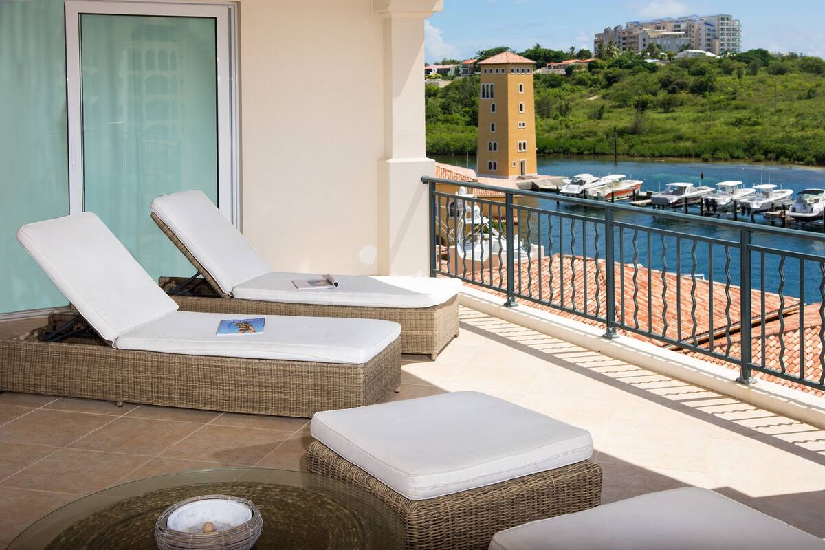 A balcony with two wicker lounge chairs and a small table, overlooking a marina with boats and a building in the background.