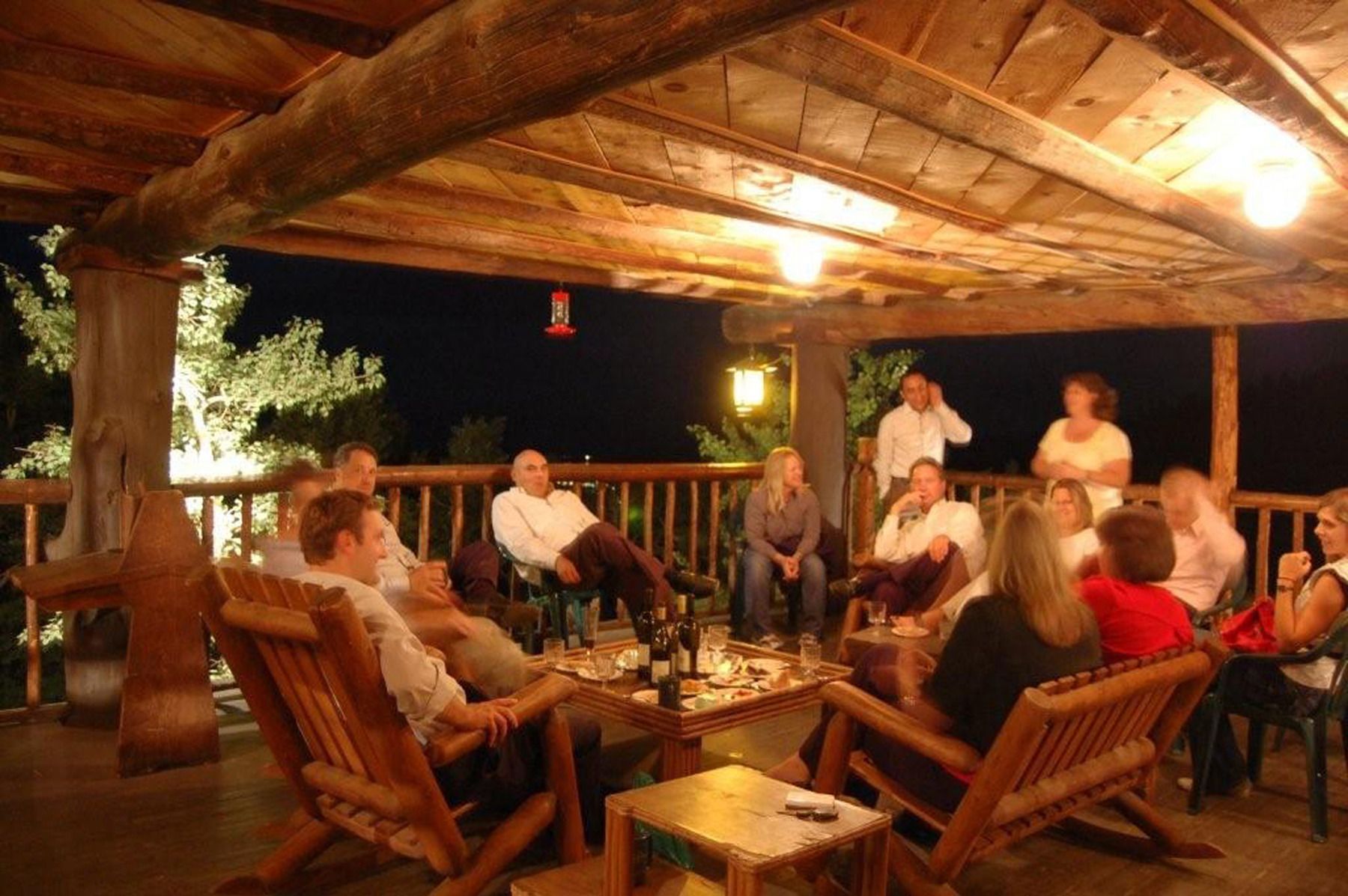 A group of people seated on wooden chairs and tables under a wooden roofed area, likely a patio or deck, with a view of trees in the background.