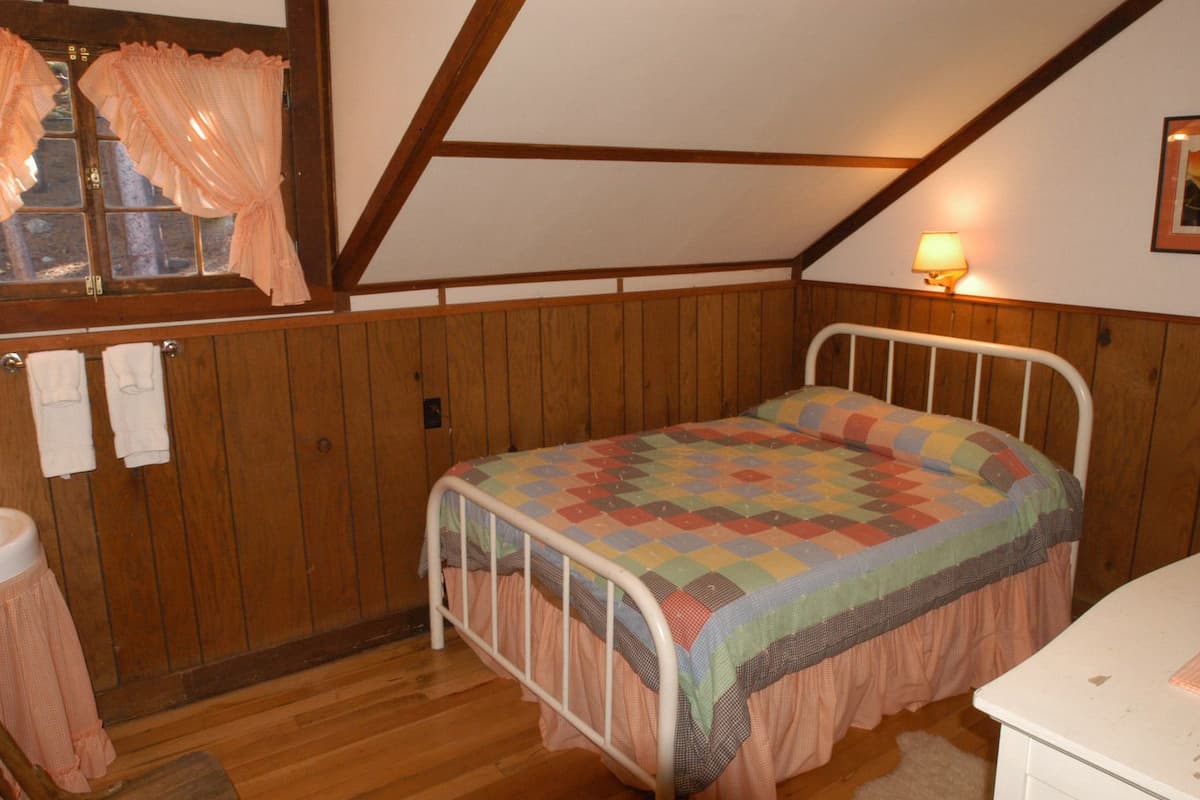 A bedroom with a wooden paneled wall, a bed with a colorful quilt, a white metal bed frame, a wooden floor, a window with curtains, a wall-mounted lamp, and a white dresser.