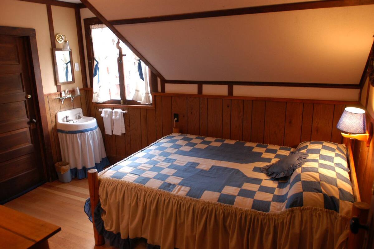 A bedroom with a checkered bedspread, wooden walls, a window with curtains, a bedside lamp, and a wooden door.
