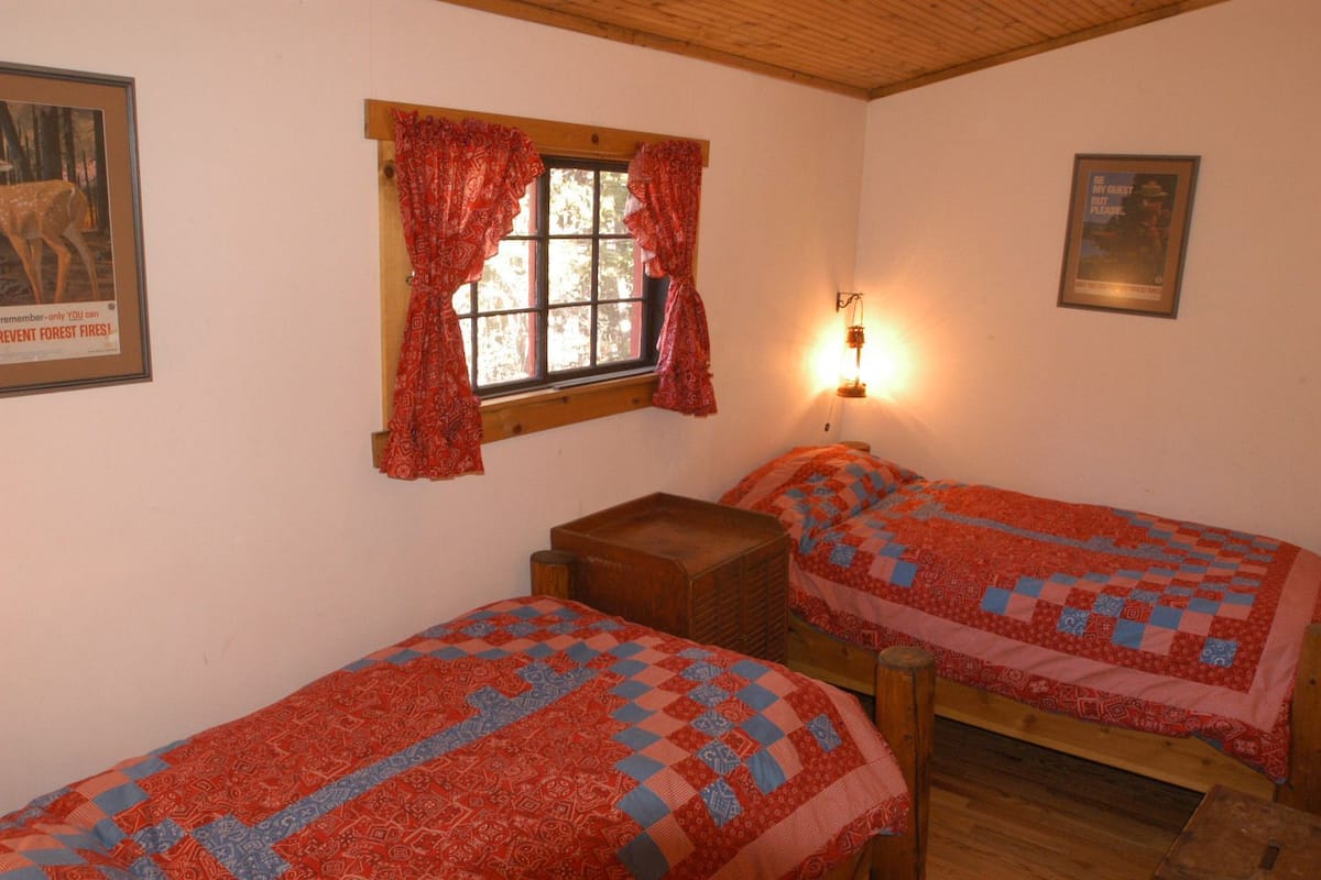 A room with two single beds, a wooden bench, a window with red curtains, and a wall-mounted light fixture.