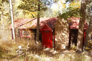 Eine rustikale Holzhütte mit roter Tür und einem Steinkamin, umgeben von Bäumen und Herbstlaub.
