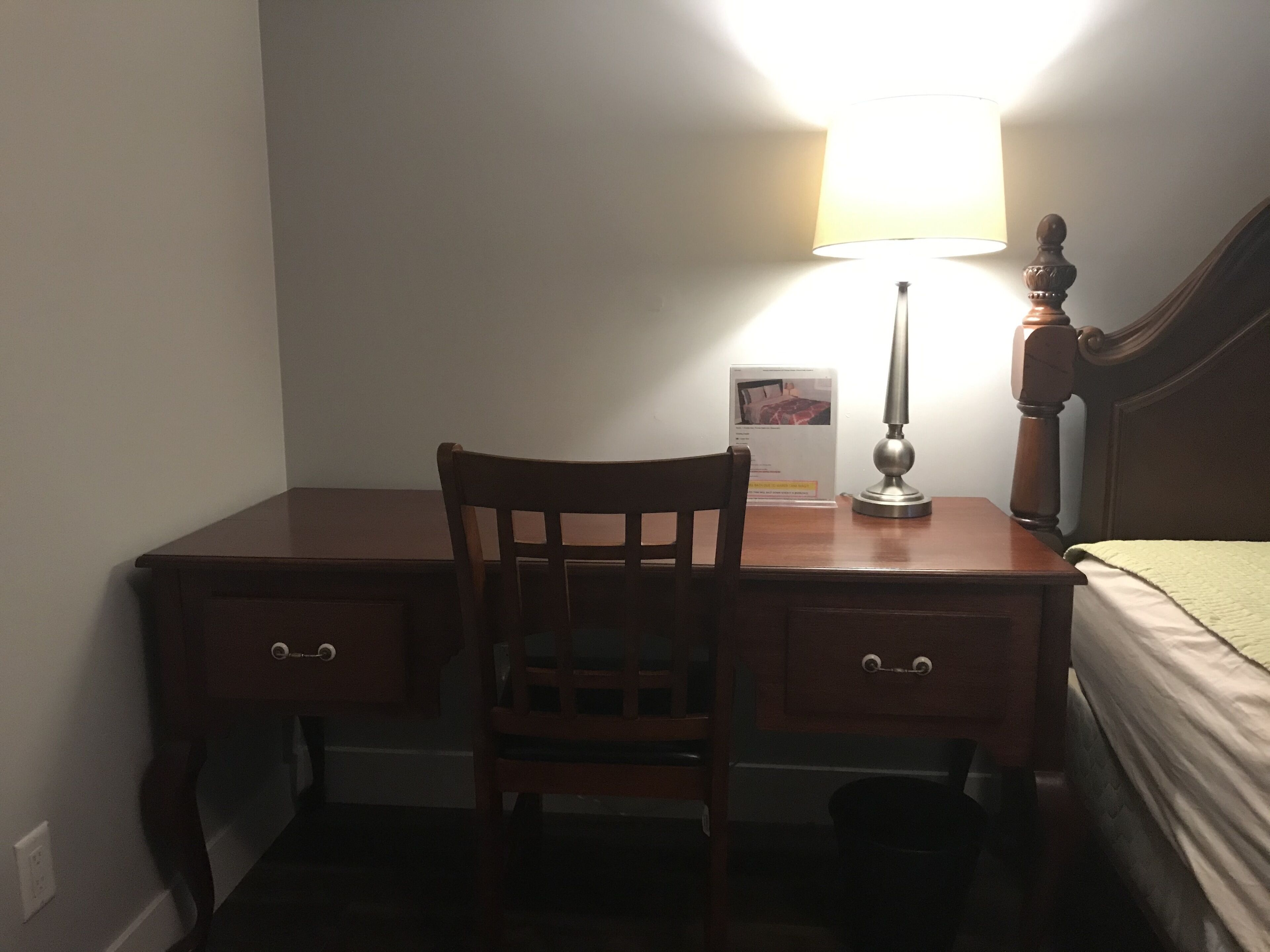Una habitación pequeña y tenuemente iluminada, con un escritorio de madera, una silla, una lámpara y una cama con una manta verde.